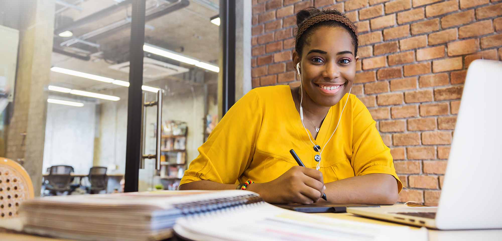 black female student wearing yellow shirt holding pen in front of laptop and books